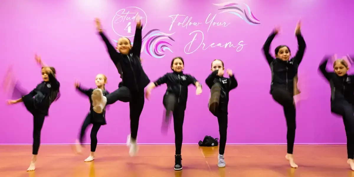 Dancers kicking in front of a purple wall with colored graphics and text that says, 