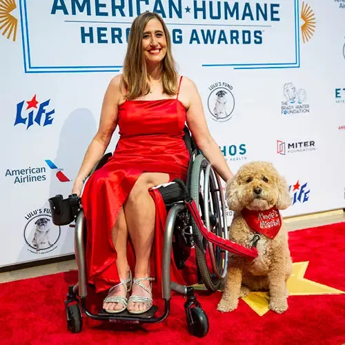 A woman in a wheelchair and her dog pose on a red carpet with gold stars in front of a step-and-repeat sign.