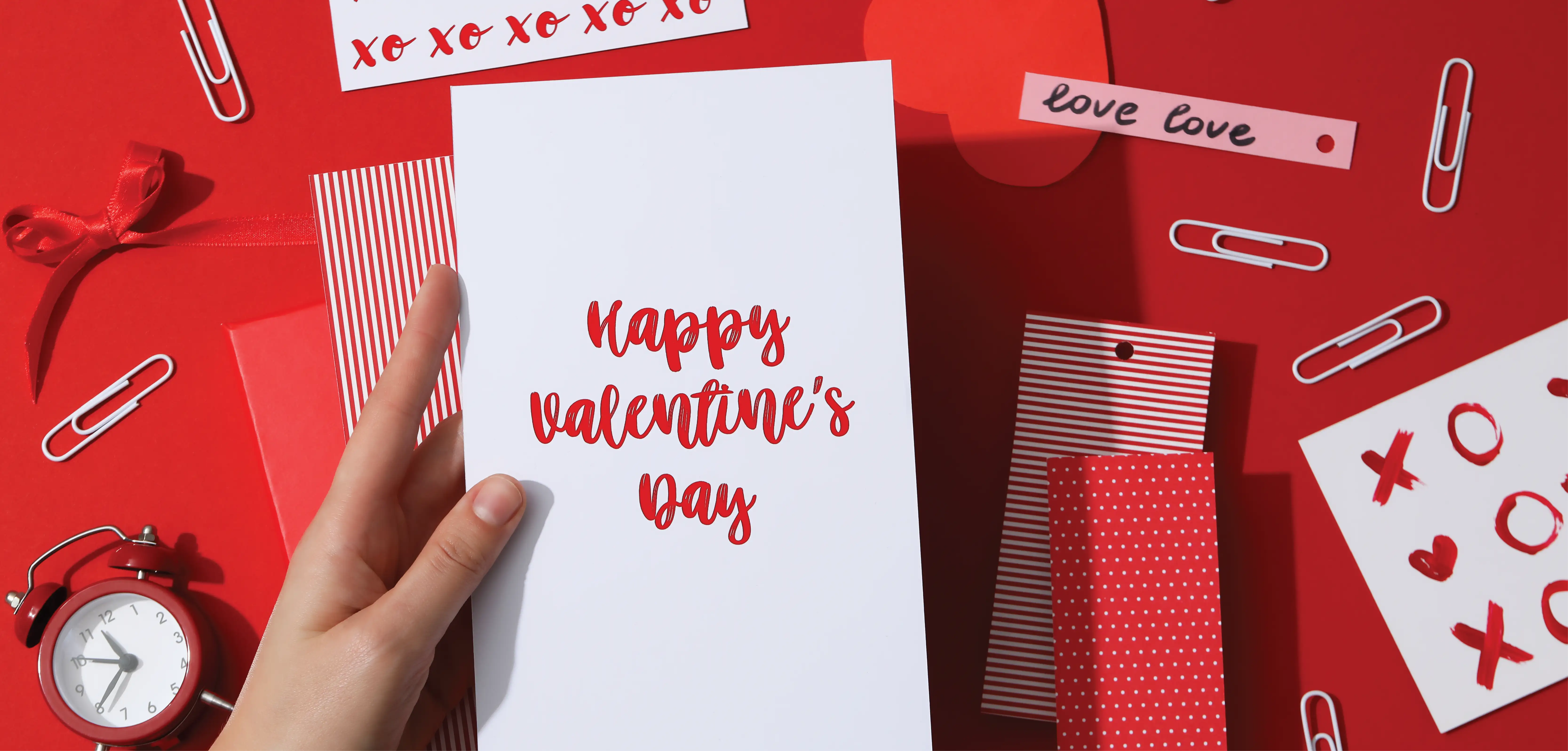 A hand holds a card that says Happy Valentine's Day above a red table scattered with paperclips and papers saying XOXO and love