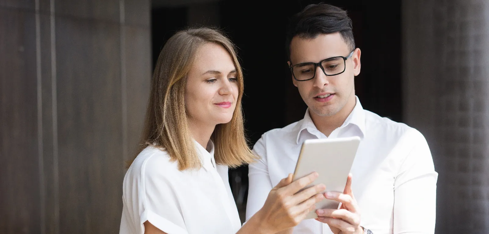 A man and woman in white collared shirts look at a tablet together