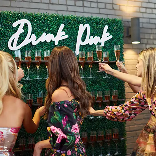 A group of women clink champagne glasses in front of a wall of greenery, holding champagne, with the words 