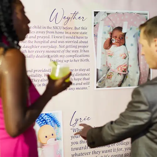 A man and woman stand in front of a sign displaying the photo, story, and name of a NICU patient